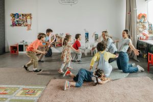 A diverse group of young children and two adults engaged in energetic play on a rug in a bright classroom, demonstrating active social interaction and shared experience. This group activity illustrates the development of social skills essential for both physical and shared digital spaces.