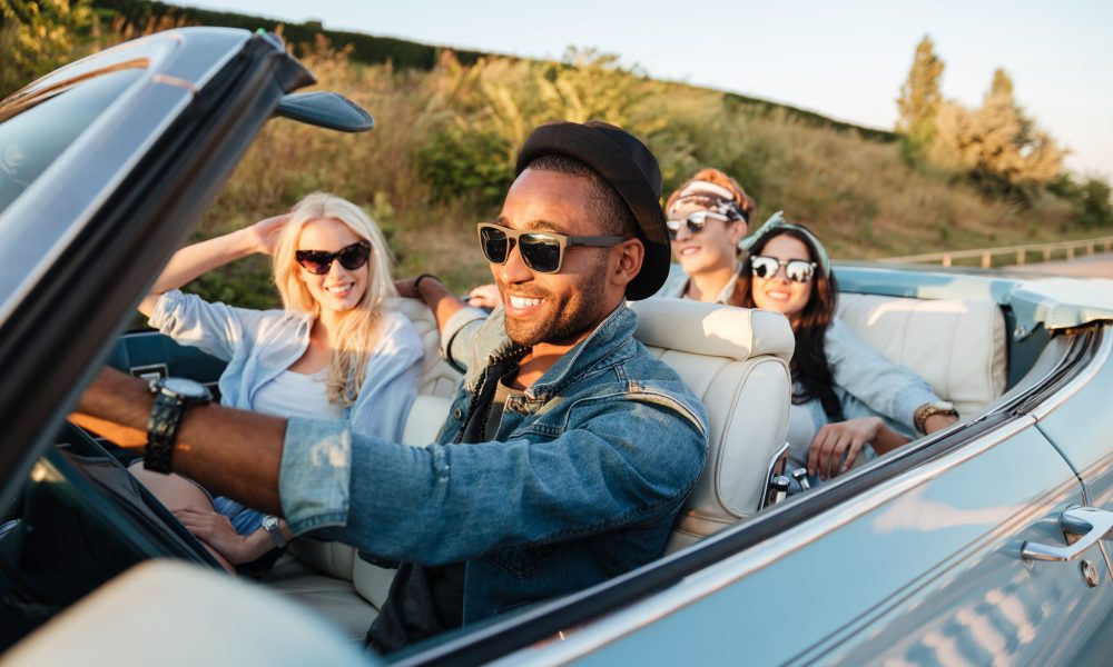 Group of cheerful young friends driving car and smiling in summer