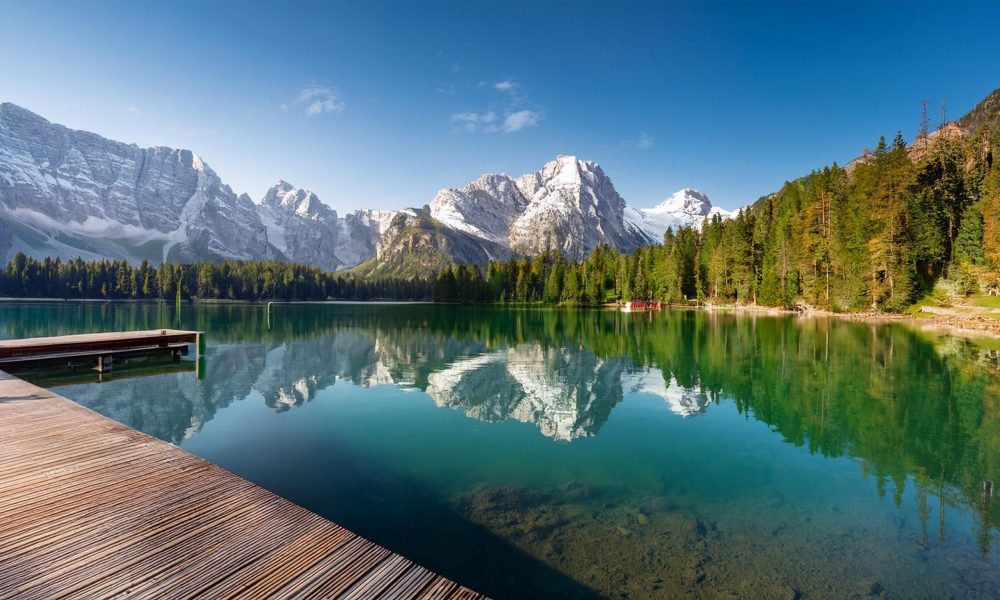 Beautiful wide shot of a lake and mountain in the background and a small deck leading to the lake, with pine trees, clouds, and snow covered mouintain tops