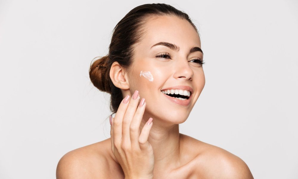 Close up beauty portrait of a laughing beautiful half naked woman applying face cream and looking away isolated over white background