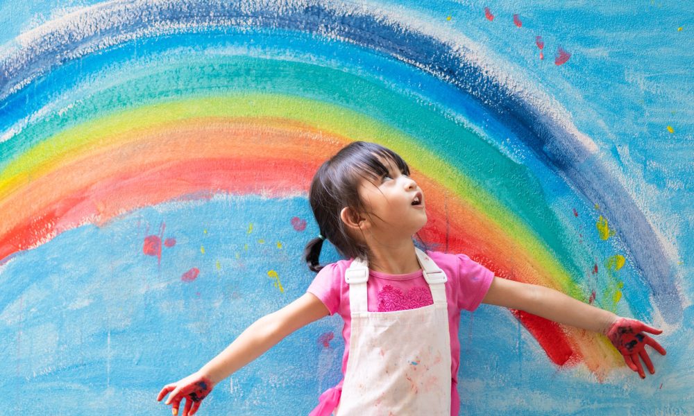 Asian little girl is painting the colorful rainbow and sky on th