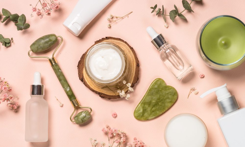 Cosmetic products - Jade roller and gua sha massager with cream and serum bottles at pastel background. Spa background. Top view with copy space.