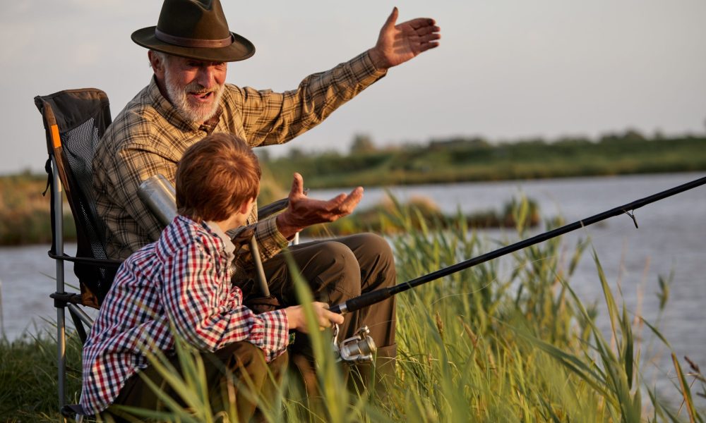 Grandfather and kid boy together fishing in the evening at sunset time in summer day on river in countryside, side view on caucasian multi-generation family, elderly man teach child boy to fish