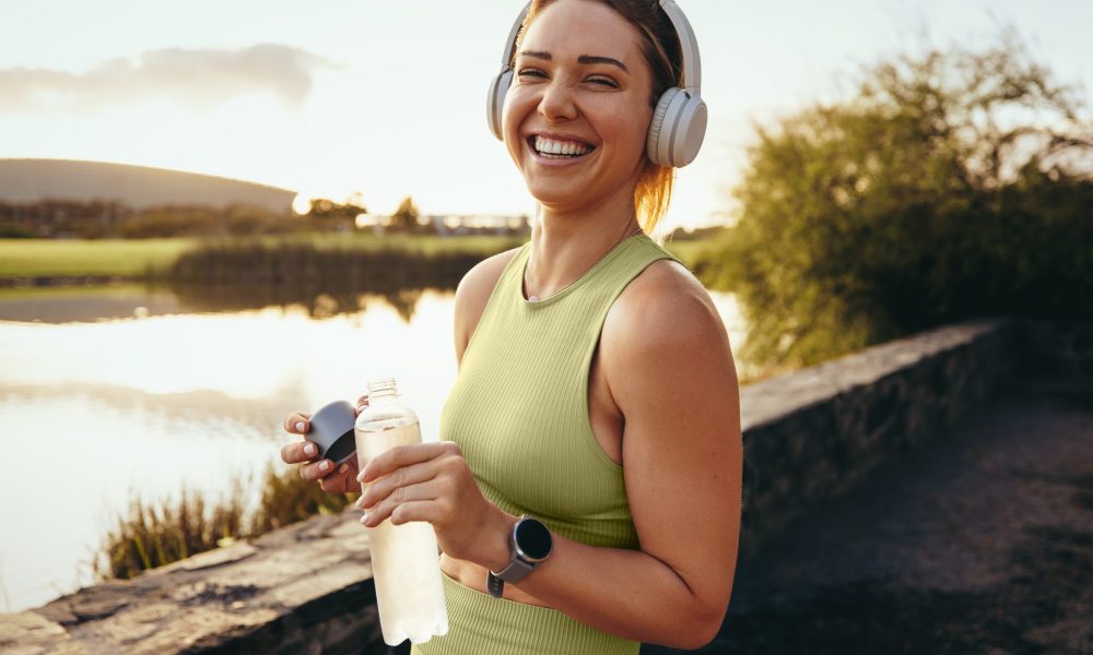 Happy sportswoman taking a water break outdoors. Fit woman standing next to a lake and looking at the camera. Woman working out with headphones in the morning.