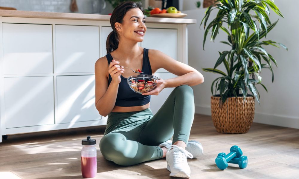 Shot of athletic woman eating a healthy bowl of muesli with fruit sitting on floor in the kitchen at home