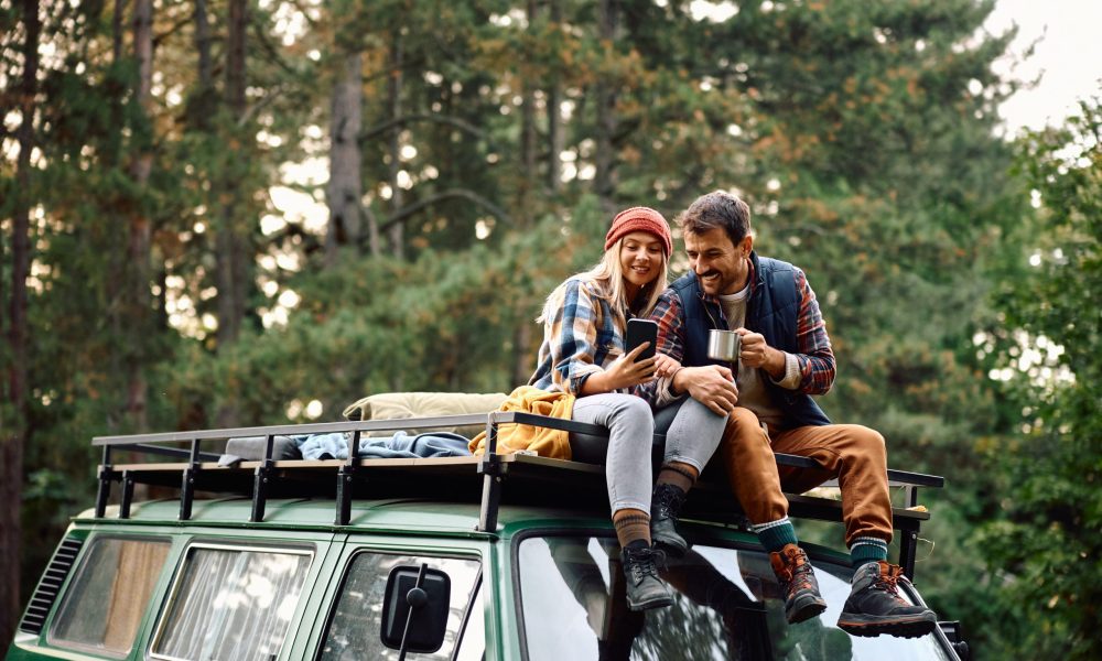 Happy couple using mobile phone while sitting on roof of their camper trailer in nature.