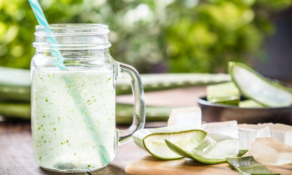 Front view of a drinking glass full of an aloe vera drink surrounded by some aloe vera crystals and sliced leaves. A black bowl full of sliced aloe vera leaves is defocused on the background. Predominant color are green and brown. Studio shot taken with Canon EOS 6D Mark II and Canon EF 24-105 mm f/4L
