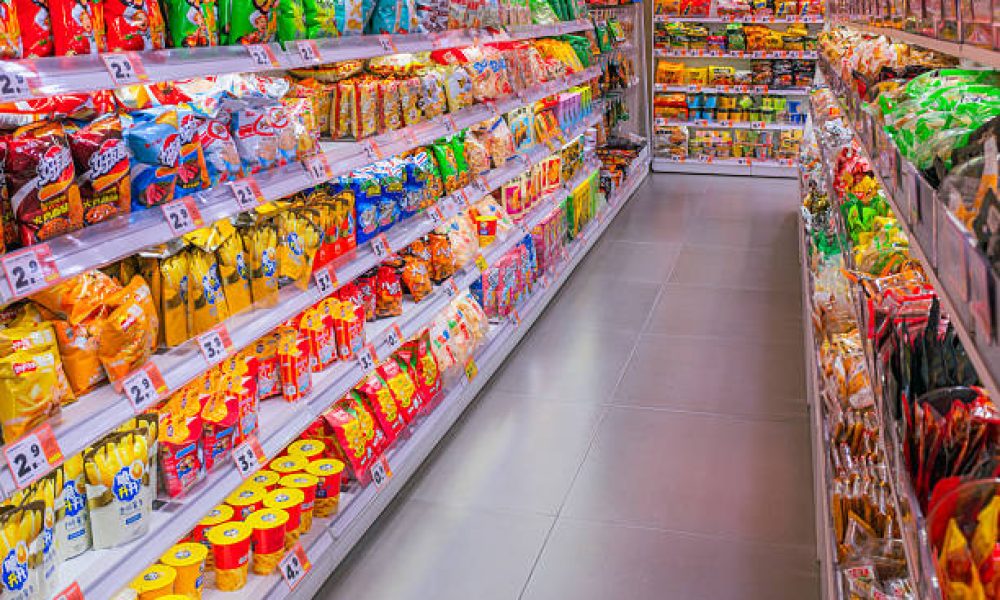 This image displays a brightly lit and colorful aisle within a supermarket or convenience store. The shelves are densely packed with a wide assortment of snack products, primarily chips and puffed snacks in brightly colored packaging. The perspective shot down the aisle creates a sense of depth and abundance, highlighting the vast selection available to customers. The clean, reflective flooring and organized shelving contribute to a modern and well-maintained retail environment.