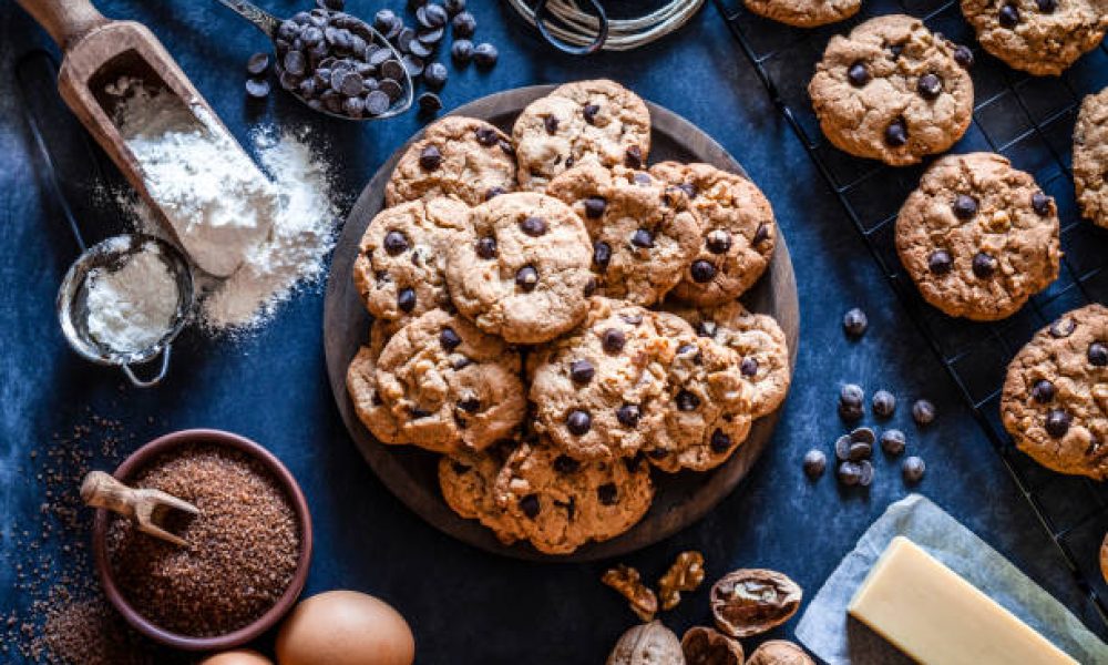 Top view of a wooden plate filled with chocolate chips cookies placed at the center of the frame. Ingredients for preparing and baking cookies are all around the plate. Some baked cookies are on a cooling rack. Ingredients includes are flour, eggs, chocolate chips, brown sugar and butter. Some kitchen utensils complete the composition. Predominant colors are brown and blue. Low key DSRL studio photo taken with Canon EOS 5D Mk II and Canon EF 100mm f/2.8L Macro IS USM