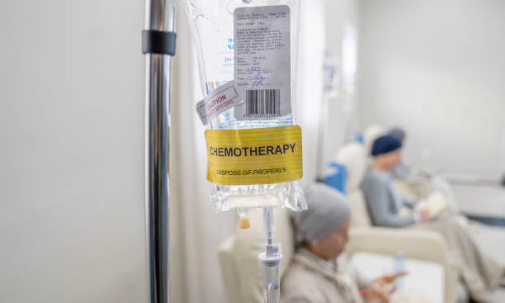 An intravenous bag of Chemotherapy hangs from an IV pole as three women sit receiving their treatment.  The women are dressed comfortably and are engaged in activities they brought to keep them busy.