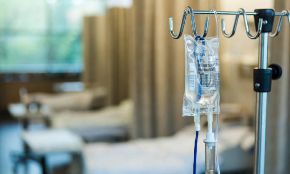The focus of the photo is on the IV bag and stand in the foreground with empty hospital beds in a row in the background.