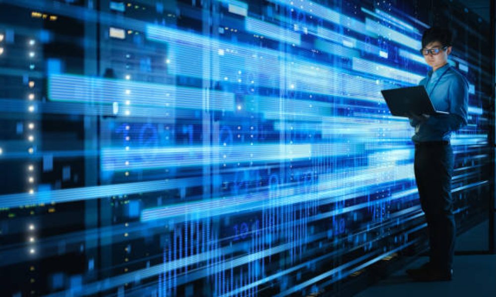 Shot of Asian IT Specialist Using Laptop in Data Center Full of Rack Servers. Concept of High Speed Internet Connections with Blue Visualization Projection of Binary Data Transfer