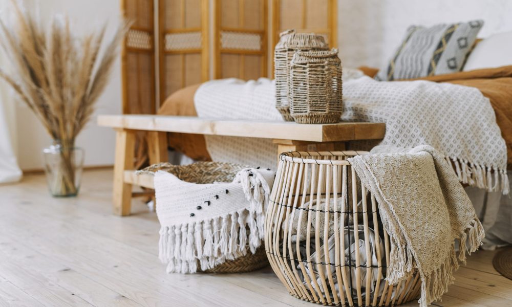 Selective focus on home decor. Comfortable bedroom in bohemian interior style with textile sheet on bed, wooden bench seat, bamboo dressing screen, dry plants in vase, wicker basket