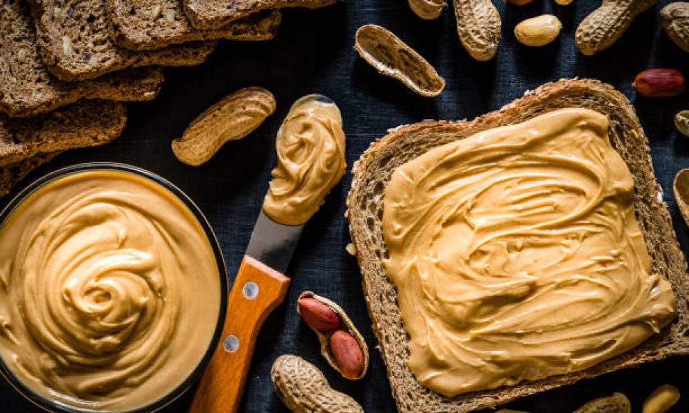 Top view of a sliced bread scattered with peanut butter on top surrounded by a kitchen knife with peanut butter on the tip, whole and peeled peanuts, some sliced breads and a bowl filled with peanut butter on a dark gray backdrop.
Low key DSLR photo taken with Canon EOS 6D Mark II and Canon EF 24-105 mm f/4L