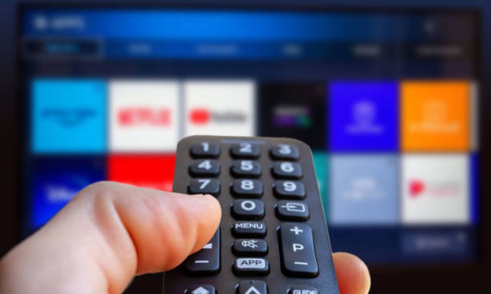 A man is holding a remote control of a smart TV in his hand. In the background you can see the television screen with streaming entertainment apps for video on demand