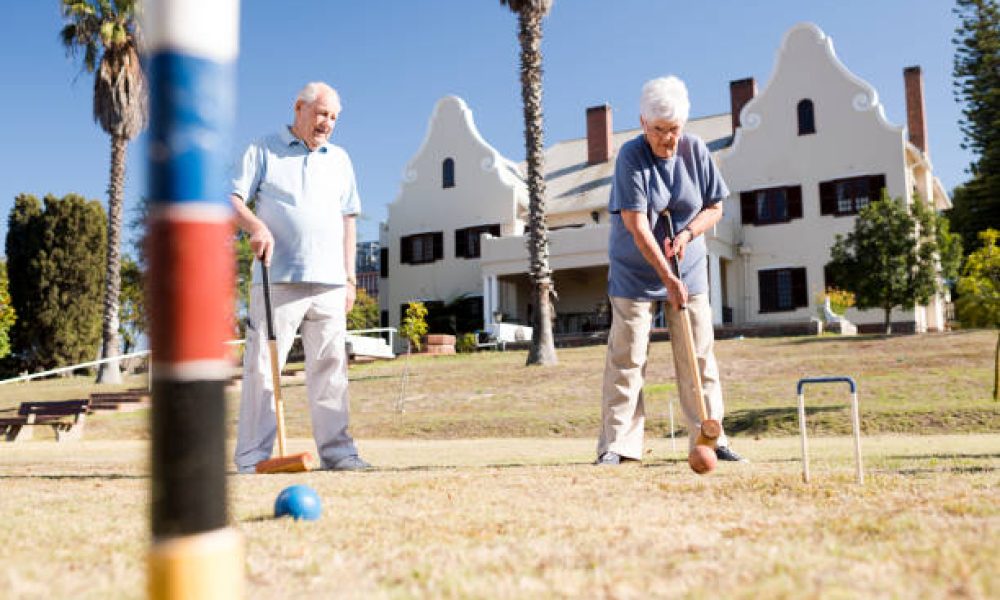 Female senior feet positioned with mallet in hand, focused on hitting the ball through the hoop, with another senior croquet player watching in the background